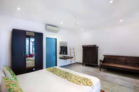 Spacious Balinese bedroom interior with white bed, wooden wardrobe, antique cabinet, ceiling fan, and tiled floor.