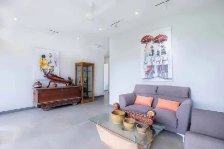 Modern Balinese-inspired living room with gray sofa, glass coffee table with wooden artifacts and ceramics, antique cabinet, and traditional wall art on white walls. - Bali Villas