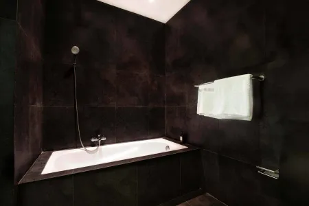 Modern black-tiled bathroom with freestanding white bathtub, handheld shower, and folded towels on rack. - Bali Villas