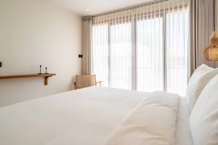 Cozy minimalist bedroom with white linens on a king bed, sheer curtains on sliding glass doors, a rattan chair, bamboo pendant light, and wooden shelf.