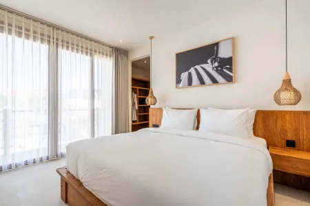 Spacious tropical bedroom with white linen bed, wooden accents, sheer curtains, rattan lamps, and abstract black-and-white wall art.