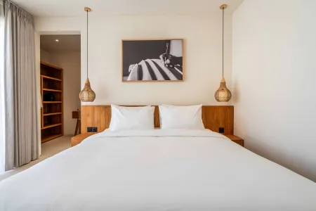 Minimalist tropical bedroom featuring a white king bed with wooden headboard, rattan pendant lights, beige curtains, and abstract black-and-white art.
