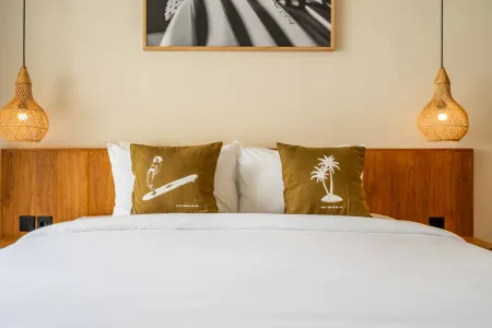 Cozy tropical bedroom with white bedding, pillows printed with a parrot on surfboard and palm tree, wooden headboard, woven pendant lights, and black-and-white surf wall art.