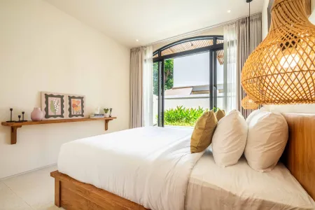 Bright, airy tropical bedroom with wooden bed, green pillows, rattan pendant lamp, and arched window overlooking lush garden.