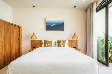 Serene tropical bedroom with king bed, white linens, palm-embroidered pillows, wooden nightstands, rattan lamps, ocean wave artwork, and sliding glass doors to lush greenery.