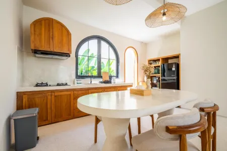 Modern tropical kitchen interior with arched wooden cabinets, white countertops, gas stove, and round dining table under rattan pendant light. - Bali Villas