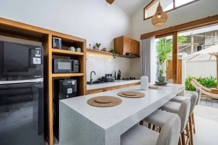 Modern tropical kitchen with white quartz island, wooden cabinets, black fridge, and open doors to lush outdoor patio. - Bali Villas