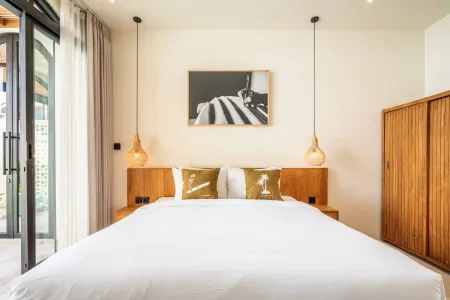 Spacious modern bedroom with white king bed, wooden headboard and wardrobe, pendant lights, abstract wall art, and sliding glass doors to lush garden.