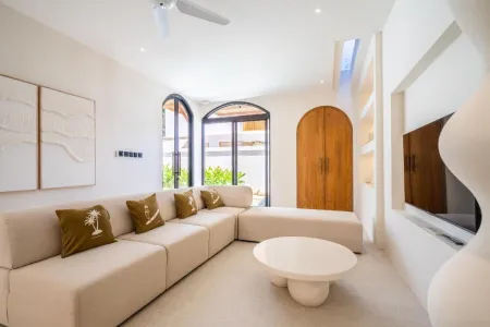 Modern minimalist living room with beige L-shaped sofa, white coffee table, abstract wall art, arched wooden doors, and tropical greenery accents. - Bali Villas