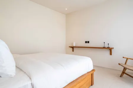 Minimalist white bedroom interior with a low wooden bed, rattan armchair, and floating wooden shelf with vase.