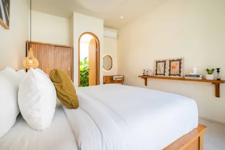Cozy minimalist bedroom with white bedding, wooden accents, hanging lantern, and archway to lush greenery.