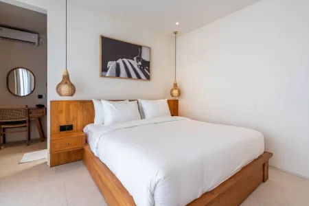 Cozy minimalist bedroom with wooden bed frame, white linens, rattan pendant lamps, and tropical wall art in a light, airy space.