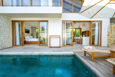 Modern tropical villa with infinity pool, wooden deck lounge chairs under umbrella, and open glass doors revealing bedroom and kitchen.