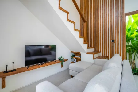 Modern living room interior with wooden staircase, wall-mounted TV on wooden shelf, gray L-shaped sofa, and tropical plants. - Bali Villas