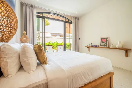 Serene tropical bedroom with white king bed, woven pendant light, and large glass doors opening to a lush balcony.