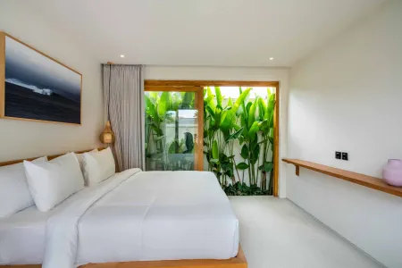 Serene minimalist bedroom with white king bed, wooden sliding doors to lush tropical greenery, and abstract seascape painting.