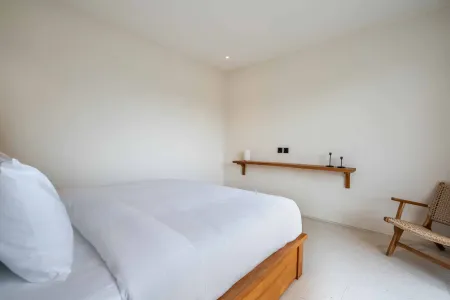 Minimalist bedroom interior with a white bed on wooden frame, floating wooden shelf with candle, and rattan armchair against neutral walls.