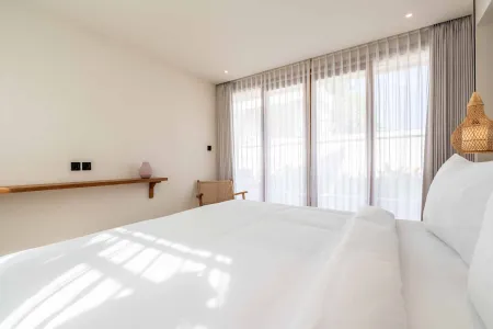Sunlit minimalist bedroom with white bed, sheer-curtained windows, rattan lamp, and wooden shelf.