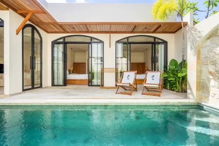 Modern tropical villa with private turquoise pool, open arched doors to a bedroom balcony, wooden lounge chairs, and lush palm greenery.