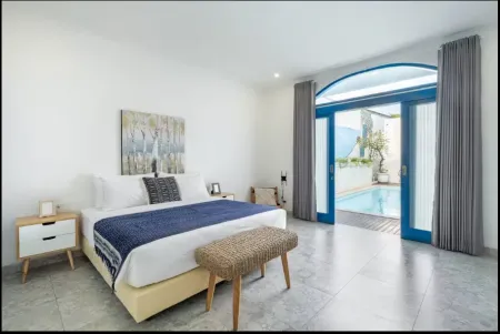 Modern bedroom with blue king bed, white nightstands, abstract wall art, rattan bench, and open sliding doors to private infinity pool.