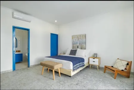 Spacious modern bedroom with king bed in navy blue bedding, wooden furniture, abstract wall art, rattan bench, and open blue door to ensuite bathroom.