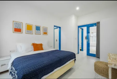 Modern minimalist bedroom with white walls, blue quilted king bed, orange pillows, abstract art frames, blue doors, and rattan seating. - Bali Villas