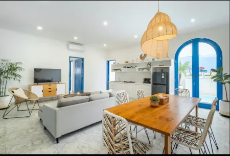 Modern open-plan beach villa interior with gray sofa, wooden dining table, rattan pendant light, and blue arches framing ocean view. - Bali Villas