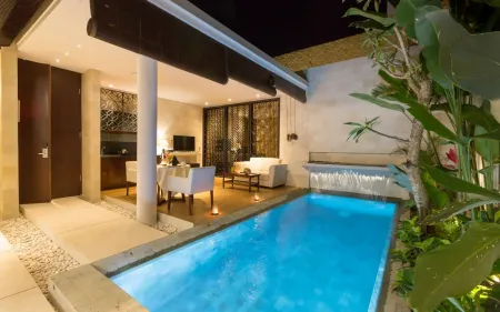 Luxurious Bali villa courtyard at night: illuminated infinity pool, outdoor dining table, sofa seating, and lush tropical plants under a wooden pavilion. - Bali Villas