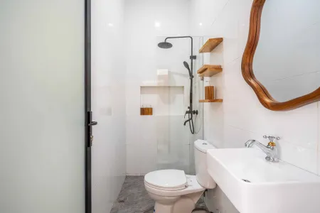 Modern compact bathroom with white tiles, glass shower enclosure, wooden shelves, white toilet, sink, and ornate oval mirror. - Bali Villas