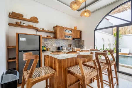 Modern tropical kitchen interior with wooden cabinets, bar stools, tiled backsplash, and arched window overlooking a pool. - Bali Villas