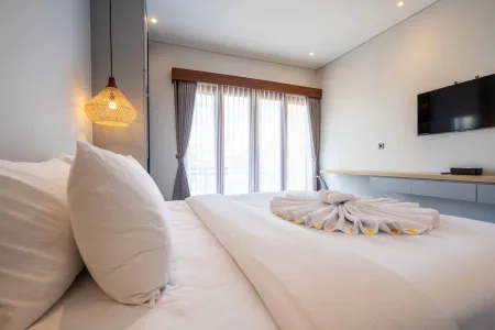 Cozy modern bedroom with white bed featuring towel flower decoration, large sheer-curtained windows, wall TV, and rattan pendant lamp.