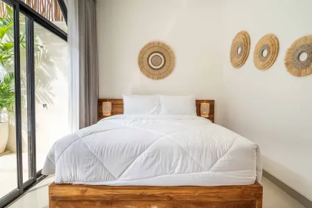 Cozy minimalist bedroom with wooden platform bed, white duvet, rattan wall hangings, and sliding doors to tropical balcony garden.