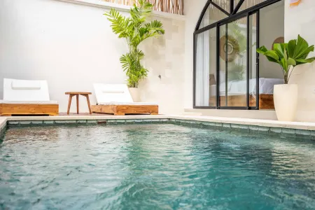 Serene tropical infinity pool in a modern villa, surrounded by white lounge chairs, lush plants, and arched glass doors to a bedroom.