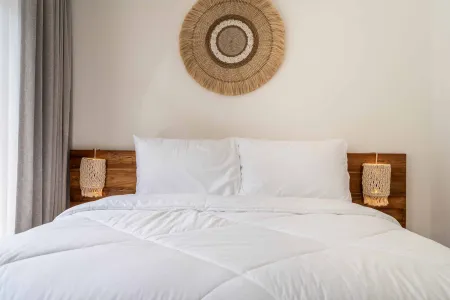 Cozy minimalist bedroom with white duvet-topped bed, wooden headboard, rattan wall decor, and boho lamps.