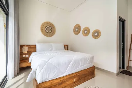 Cozy tropical bedroom with wooden platform bed, white bedding, rattan wall art, and sheer-curtained window.