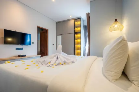 Spacious luxury hotel bedroom with king-size white bed, folded towel, yellow petals, wall-mounted TV, and wooden wardrobe.
