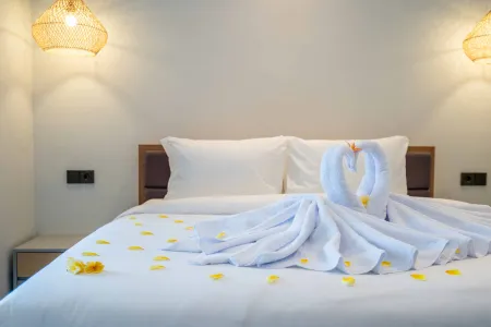 Romantic white swan towel art forming a heart on a neatly made bed, surrounded by yellow flower petals, in a softly lit minimalist bedroom.