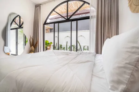 Cozy tropical bedroom with white bedding, arched black-framed window overlooking potted succulents, and leaf-shaped mirror.