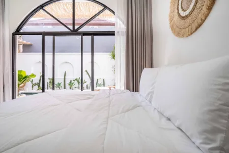 Cozy tropical bedroom with white duvet bed, woven rattan wall hanging, and arched window overlooking lush green courtyard garden.