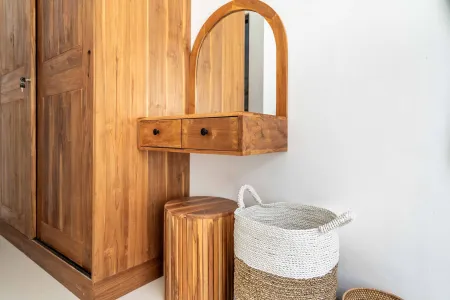 Wooden arched vanity mirror with drawers beside a tall teak wardrobe, a slatted stool, and woven baskets in a minimalist bedroom.