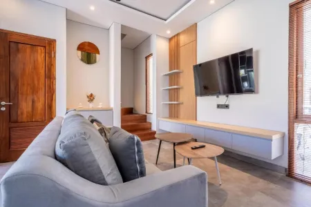 Modern living room interior with gray sofa, wooden coffee table, wall-mounted TV, floating shelves, orange mirror, and stairs in a tropical-style home. - Bali Villas