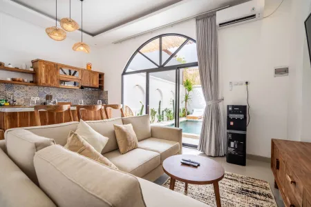 Spacious tropical villa living room with beige sofa, wooden coffee table, open kitchen bar, and arched window overlooking private pool. - Bali Villas