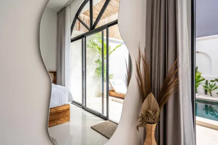 Modern bedroom with wavy wall mirror reflecting arched sliding doors to lush poolside patio and plants.