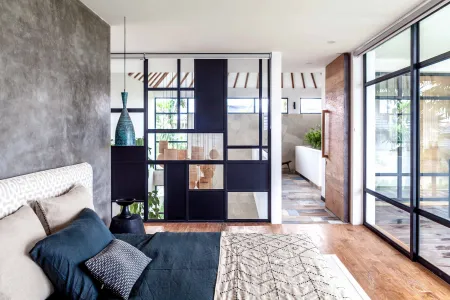 Modern bedroom interior with teal bedding on a platform bed, sliding glass partition, wooden floors, and lush greenery beyond large windows.