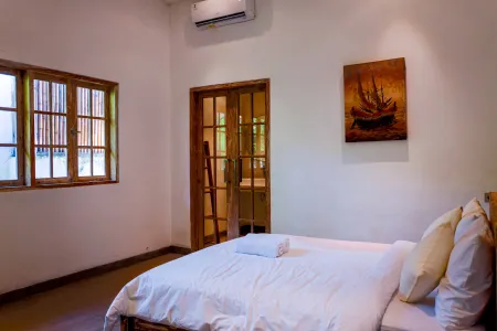 Cozy tropical bedroom with white linens on wooden bed, nautical ship painting, louvered windows, and sliding doors to en-suite bathroom.