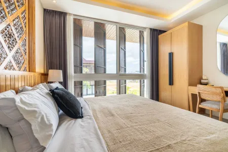 Spacious tropical bedroom with king bed, wooden wardrobe, desk, and large sliding windows overlooking lush greenery.