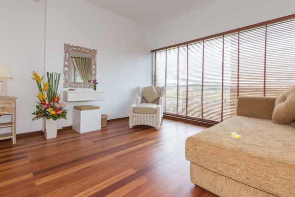 Elegant Balinese bedroom interior with white vanity, tropical flower arrangements, beige armchair, and large wooden-shuttered windows overlooking lush greenery.