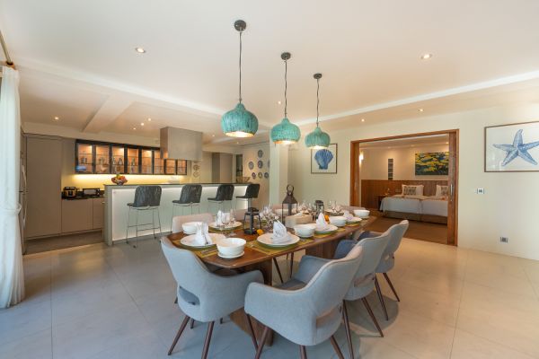 Modern open-plan dining room in a tropical villa, featuring a wooden table set with white linens and turquoise pendant lights, adjacent to sleek kitchen and bedroom.