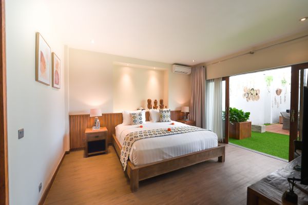 Spacious tropical bedroom with wooden king bed, sheer curtains, potted plants, and open doors to a lush green garden.