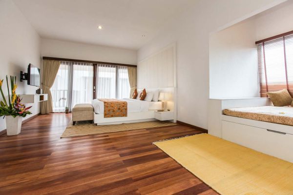 Spacious Balinese-style luxury bedroom with king bed, wooden floors, large curtained windows to balcony, potted tropical plants, and corner daybed.